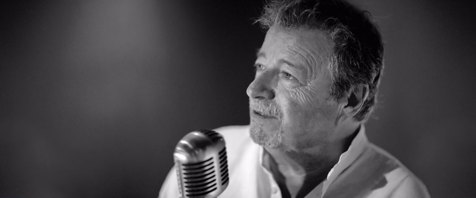 Portrait noir et blanc de Jean-Claude Donda, comédien voix off Sonacom, en chemise claire, le regard levé, au micro vintage