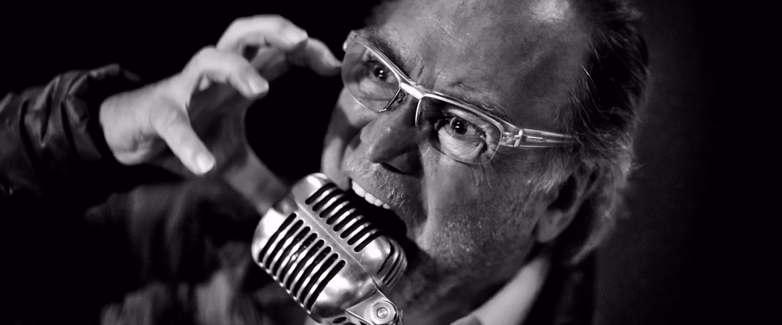 Portrait noir et blanc de Michel Elias, comédien voix off Sonacom, à lunettes, la main levée, au micro vintage