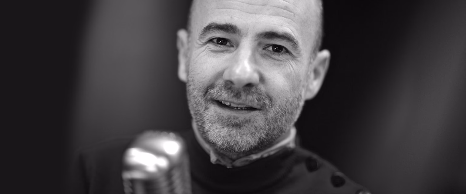 Portrait noir et blanc de Patrick Kuban, comédien voix off Sonacom, souriant, au micro vintage
